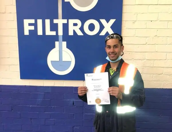 Ben, stood indoor in front of a FILTROX sign, showing his apprenticeship certificate